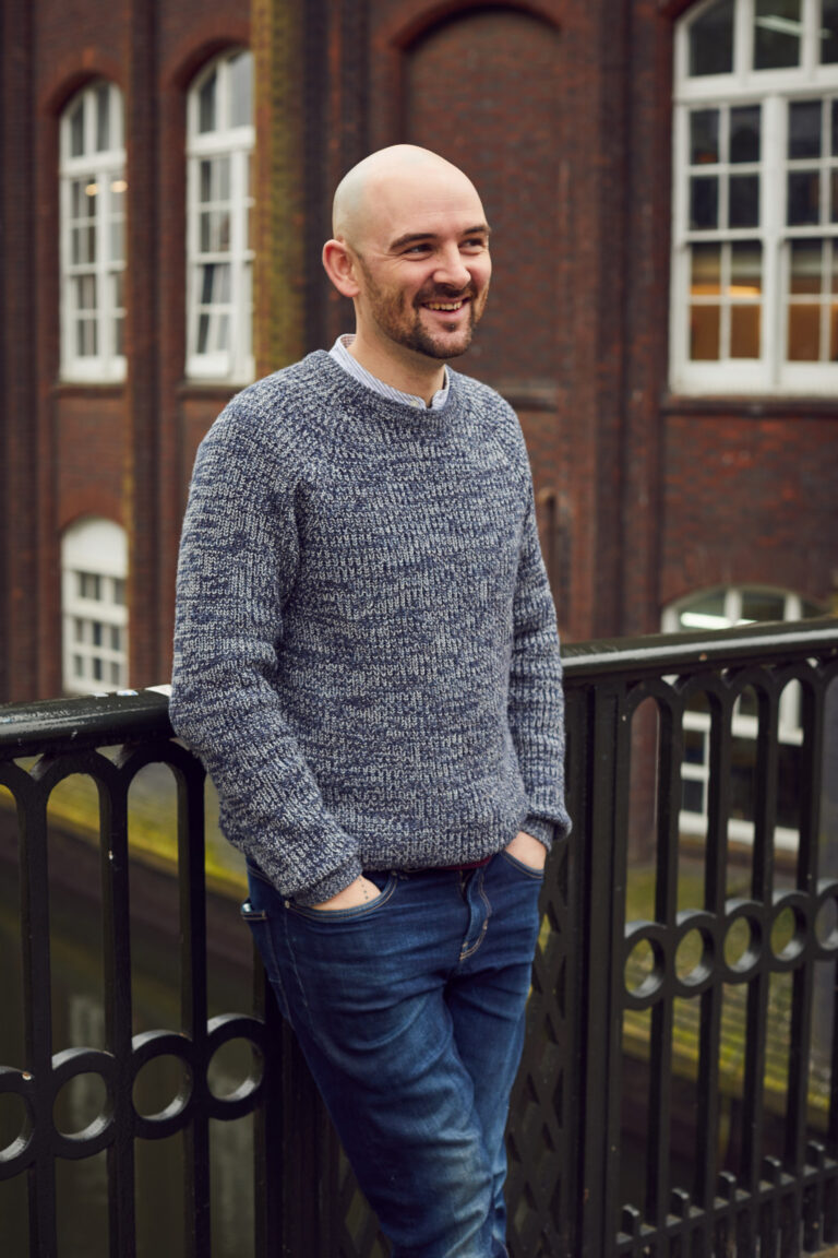 Graphics Lecturer at Norwich University of the Arts, Jamie Johnstone standing on the St Georges bridge in Norwich wearing jeans and a jumper