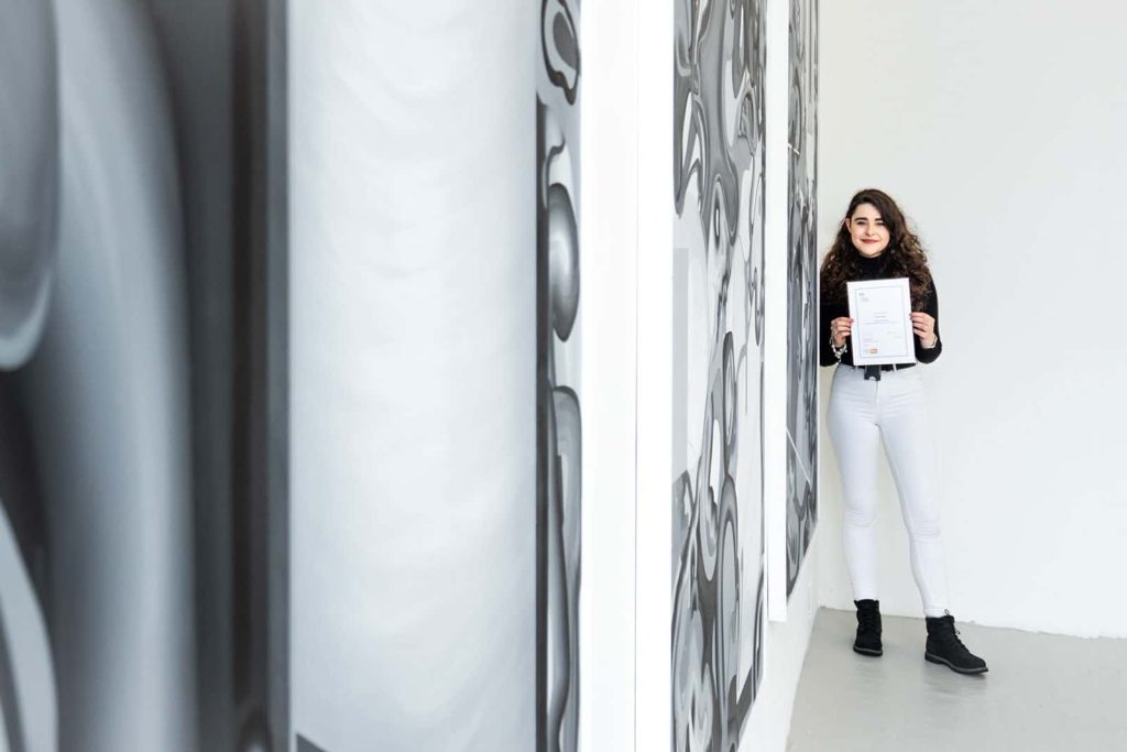 Aurellia Coster, BA Fine Art graduate standing next to her grey and white work holding her Jennifer Davey Award Certificate