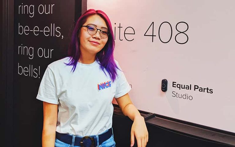 Young woman in a white t-shirt and pink and purple hair leaning against a wall and smiling at the camera