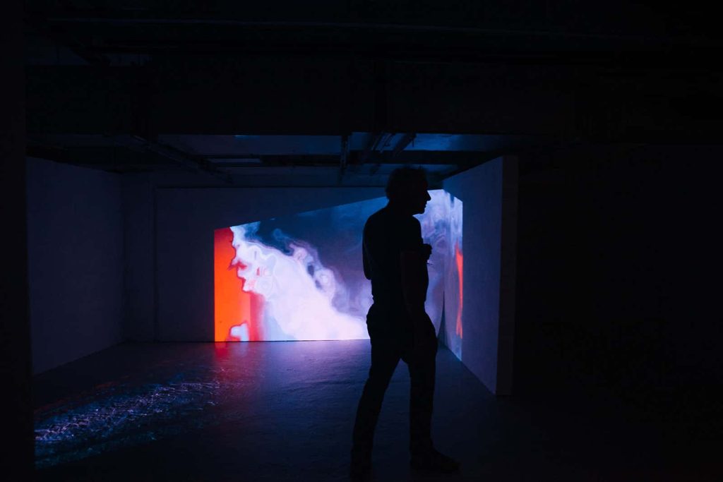 Atmospheric photo of a man walking through a dark room in front of a screen with a colourful film on at the Norwich University of the Arts MA Degree Shows