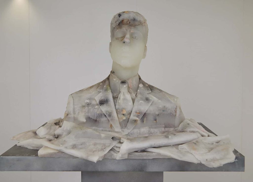 Photo of a wax bust of a man in a suit and tie on a grey plinth