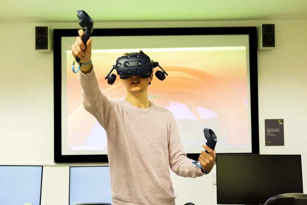 A man using the VR Lab at Norwich University of the Arts, wearing a VR headset lifting his arms up in front of a projector