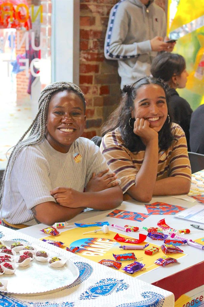 Two members of a Norwich University of the Arts Students Union Society sit at the SU fair smiling