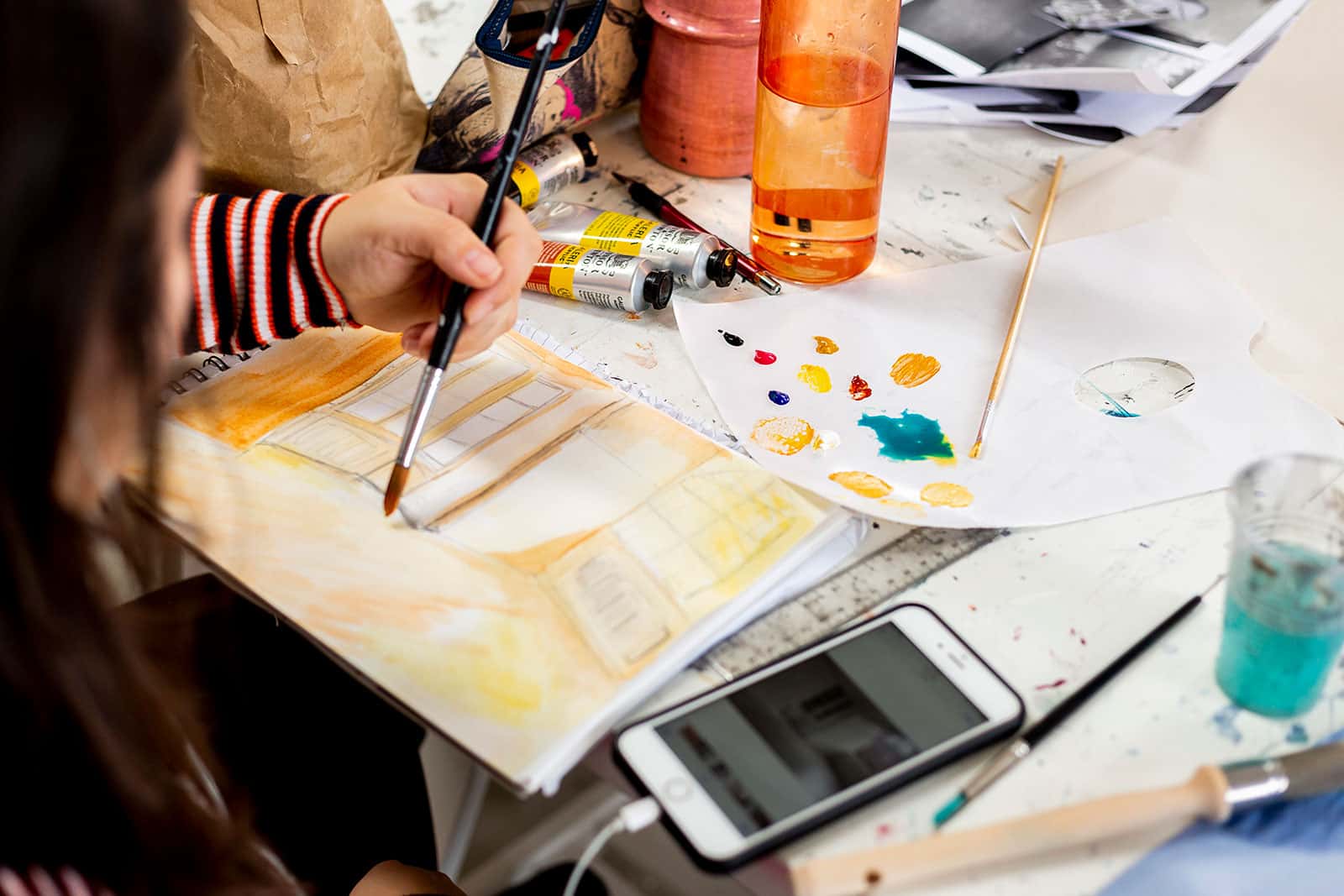 A Student holding a paint brush at Norwich University of the Arts, painting on a sketch book with a phone next to her surrounded by oil paints and a water bottle