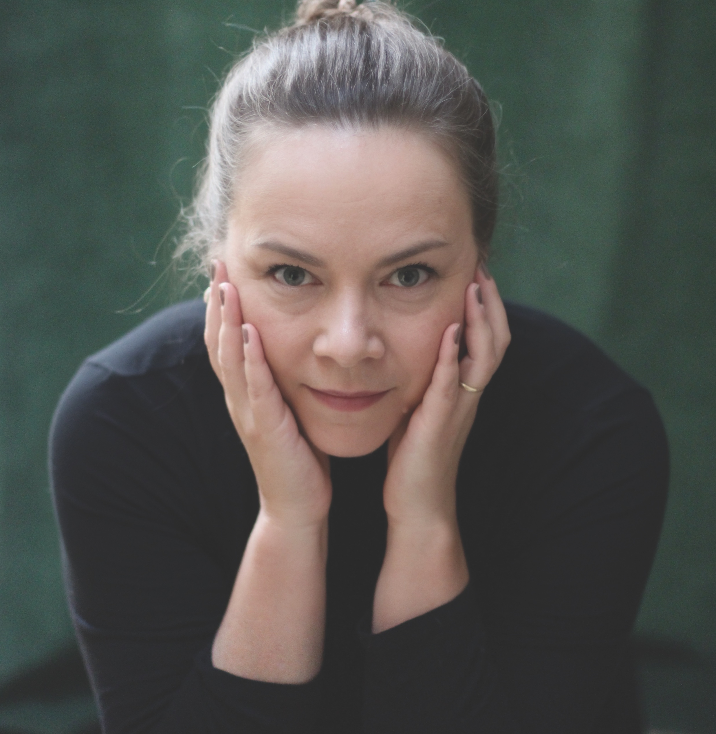 A woman with hair in a bun rests her face on her hands, looking directly at the camera with a calm expression, against a blurred dark green background. She wears a black top and a simple ring.
