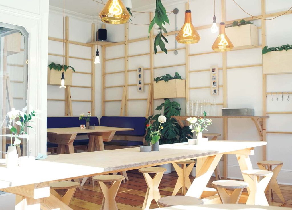 The interior of a cafe in lots of natural light, with pale wood tables and shelves dotted with plants