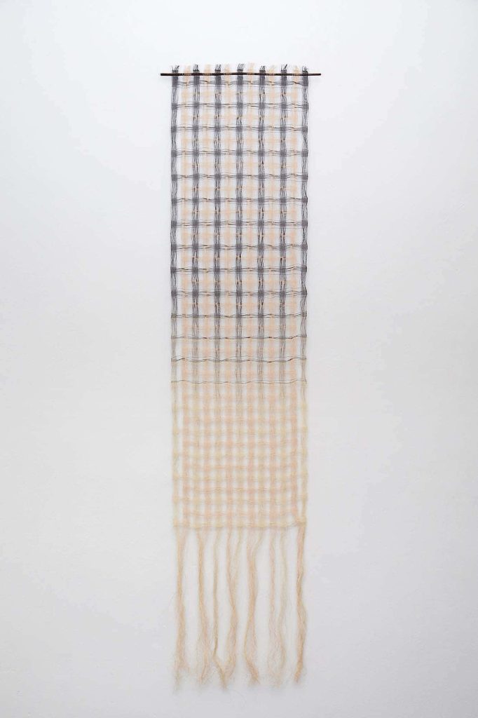 Long rectangular textiles piece hanging on a white wall, made of woven squares in cream and blue