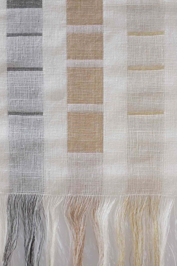 Close up of a woven textiles piece hanging on a white wall and made up of grey and pale rust squares on a cream background