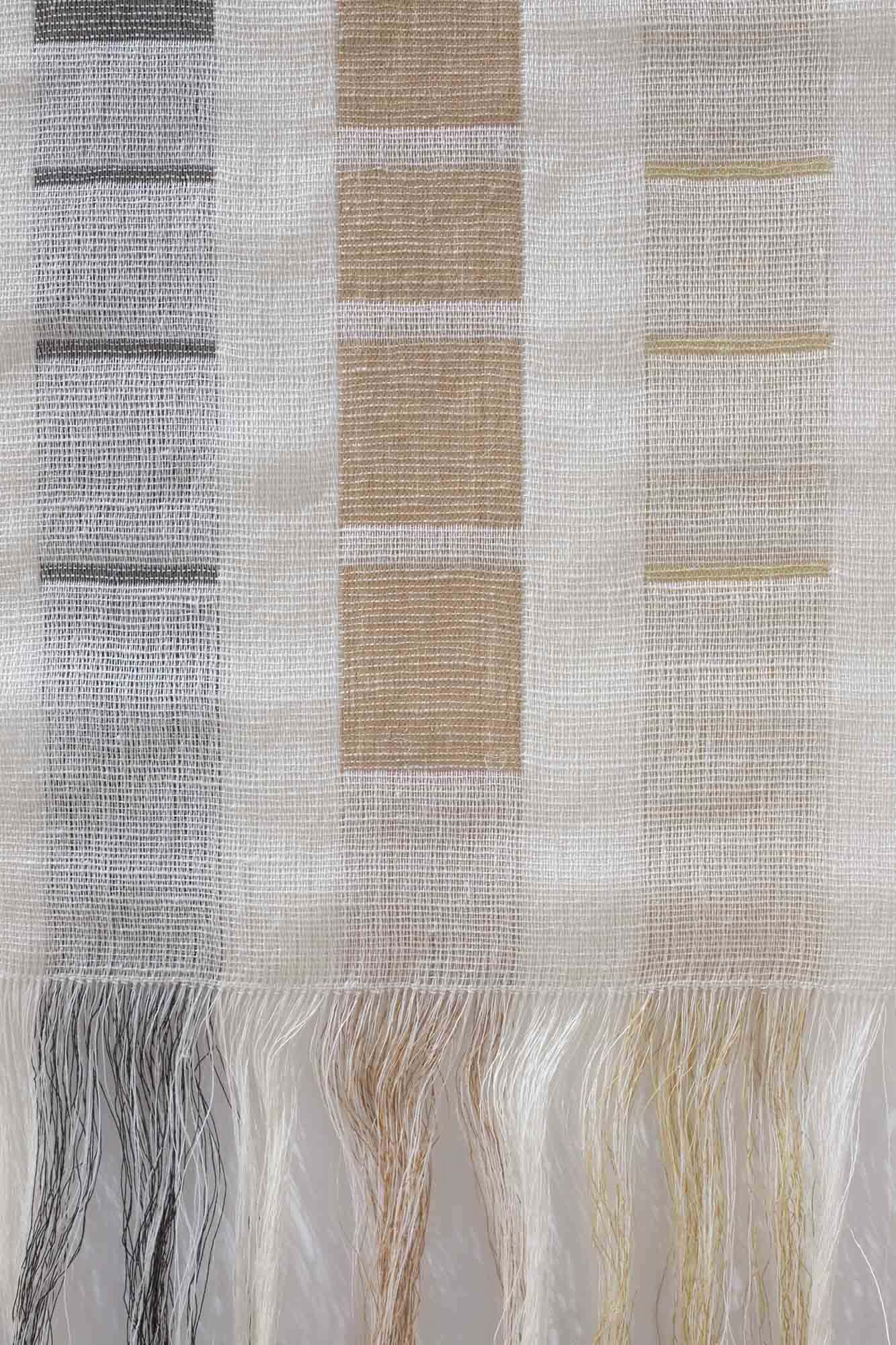 Close up of a woven textiles piece hanging on a white wall and made up of grey and pale rust squares on a cream background