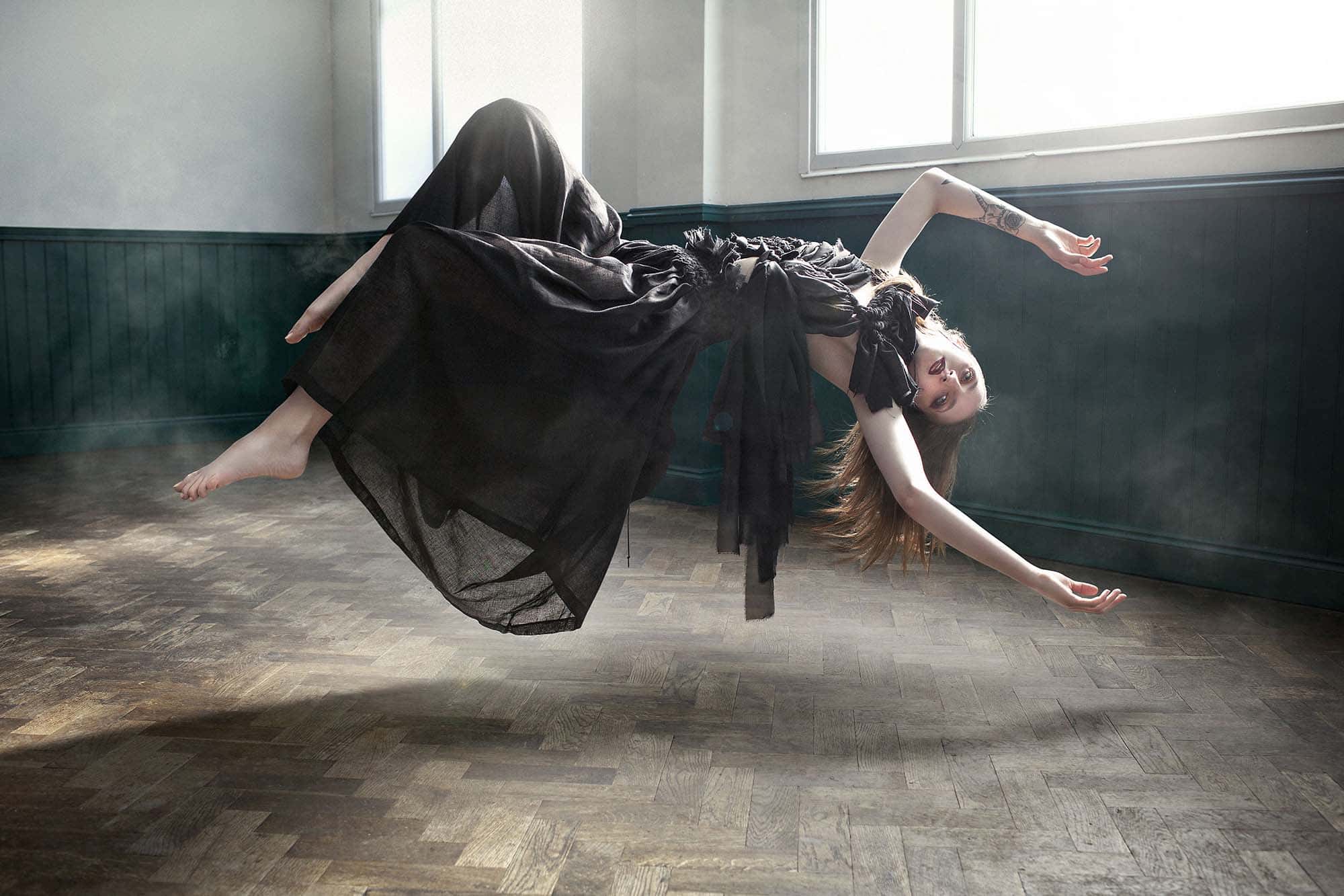 Photo by Kev Foster showing a woman in a black dress levitating horizontally with her arms above her head in soft natural light