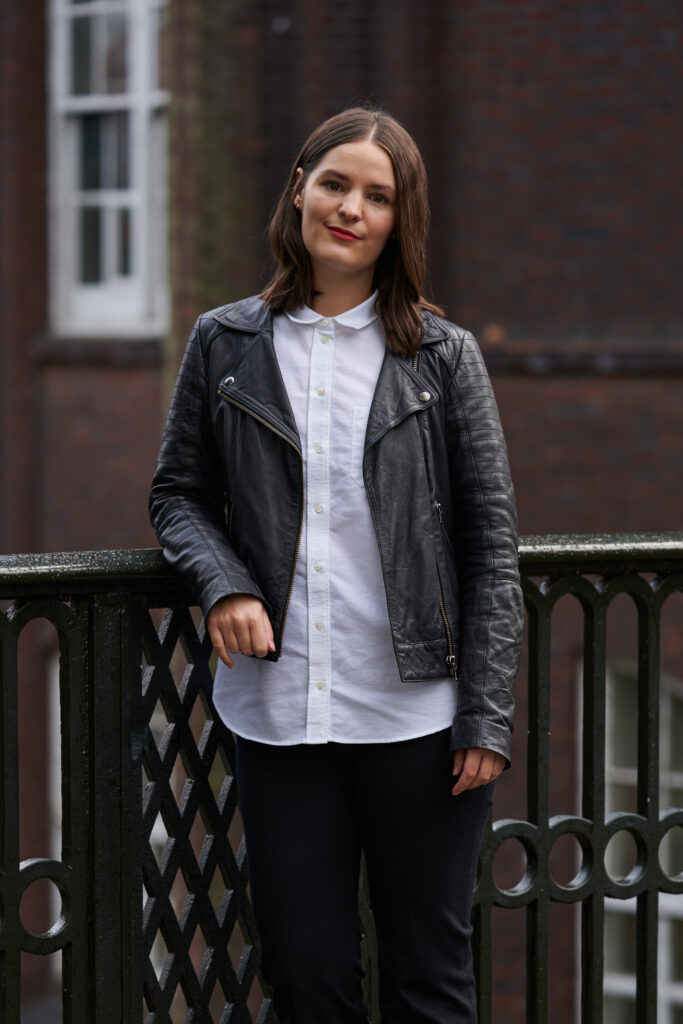 BA Fashion Marketing and Business Lecturer Holly Farrar stands for a profile photo in a leather jacket and white shirt, smiling at the camera