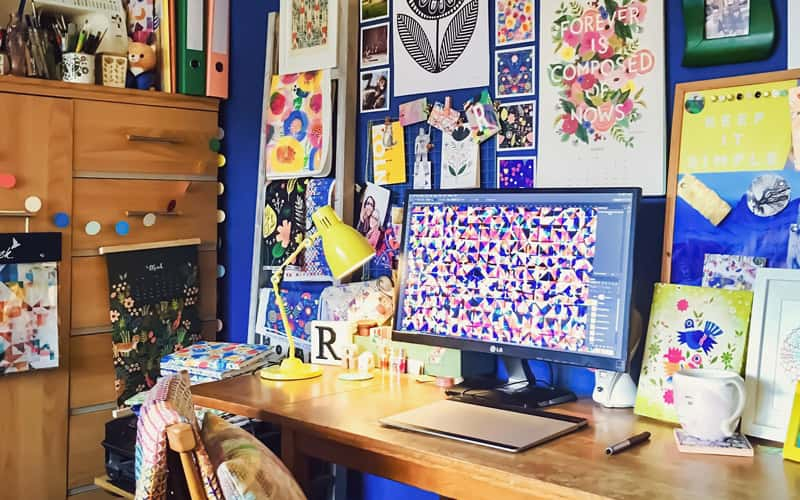 BA Textile Design graduate Rachel Parker's home studio. Her wooden desk, with graphics tablet and monitor is up against a bright cobalt blue wall. On the wall is a gallery of colourful prints, calendars and embroidery, some created by Rachel.