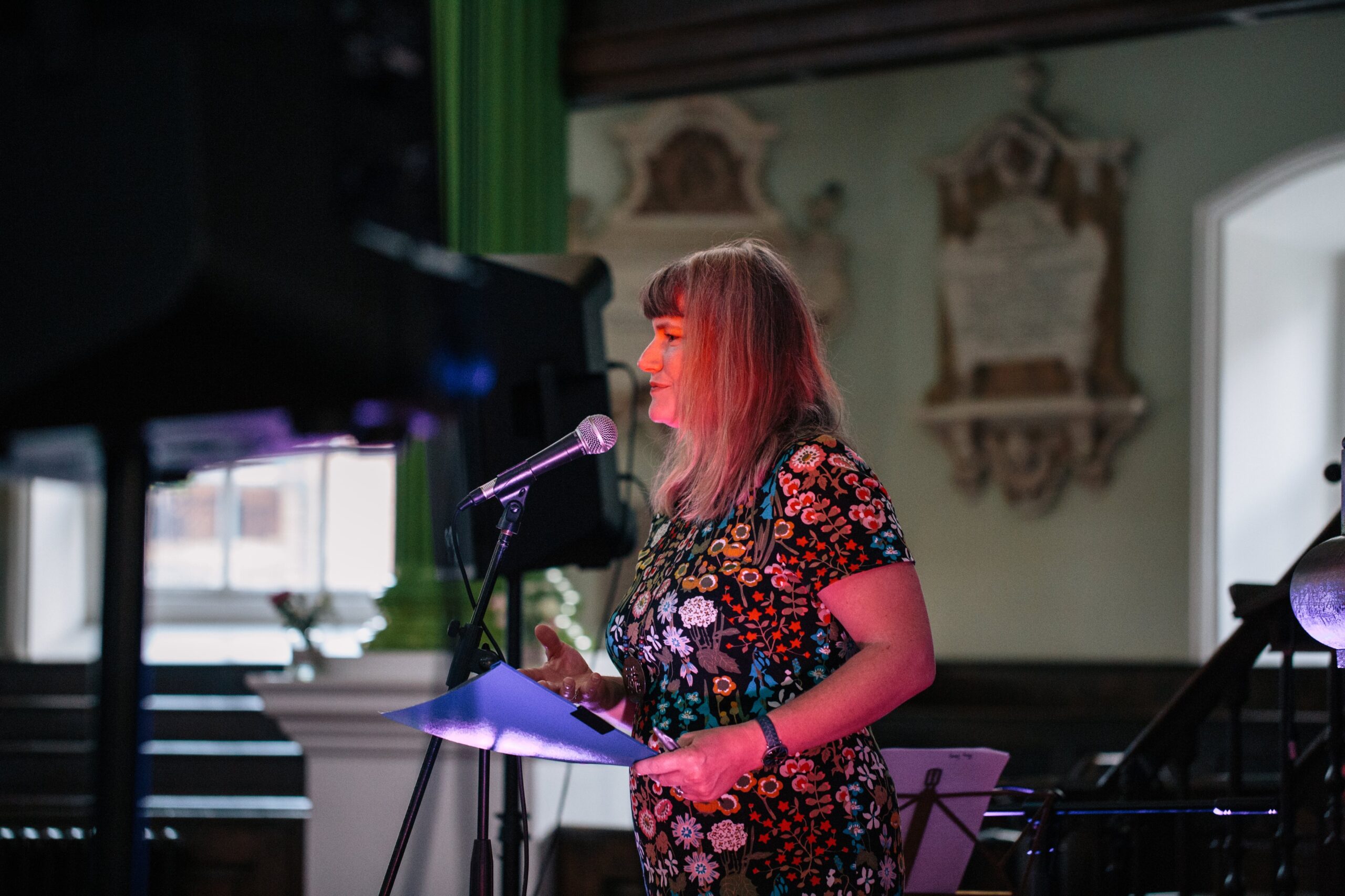 A woman with long hair, wearing a colorful floral dress, speaks into a microphone in a well-lit room with green columns and decorative wall plaques in the background. She holds papers and appears to be giving a presentation.