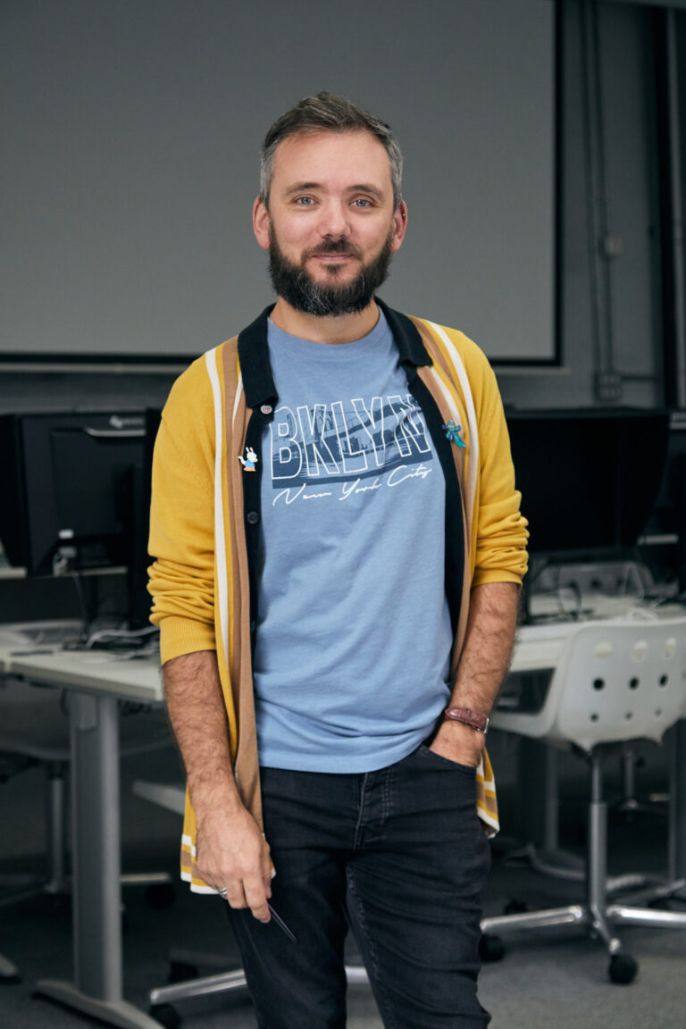 A bearded man stands in a classroom, wearing a blue "BKLYN New York City" t-shirt, a yellow cardigan, and black jeans. He poses with a relaxed expression, hands in his pockets, surrounded by desks and computer monitors.