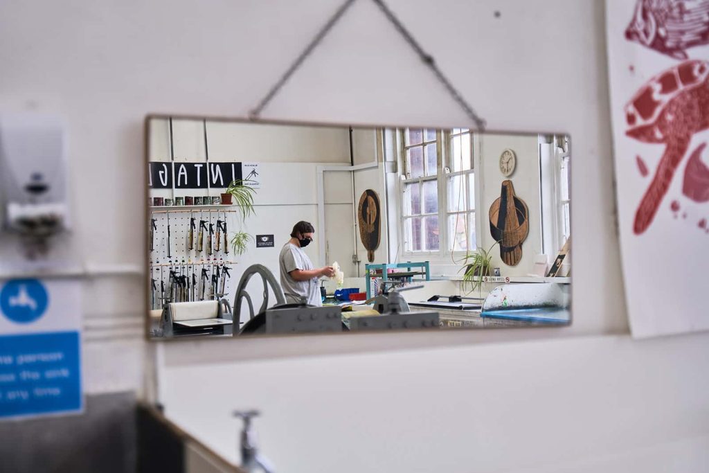 A mirror showing a reflection of a person working in a studio with a mask on at Norwich University of the Arts