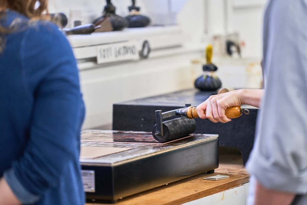 Hand rolling ink on a print with someone watching in the Norwich University of the Arts Printmaking Workshop