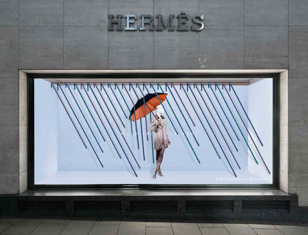 Store window visual merchandising for Hermes, featuring a sky blue backdrop, with rain falling. In the middle is a mannequin wearing a Hermes coat and holding an umbrella up. By BA Fashion Communication and Promotion student Beth Poulter