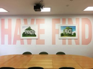 wooden table in white room. big pink words on wall saying "have find' and two artworks infont saying strength and peace