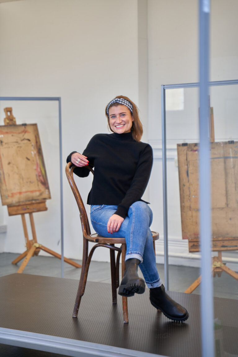 Claire allergen sitting on wooden chair with legs crossed and one arm on the back of chair on black platform. Claire has jeans and black jumper on. 2 artist easels in background.