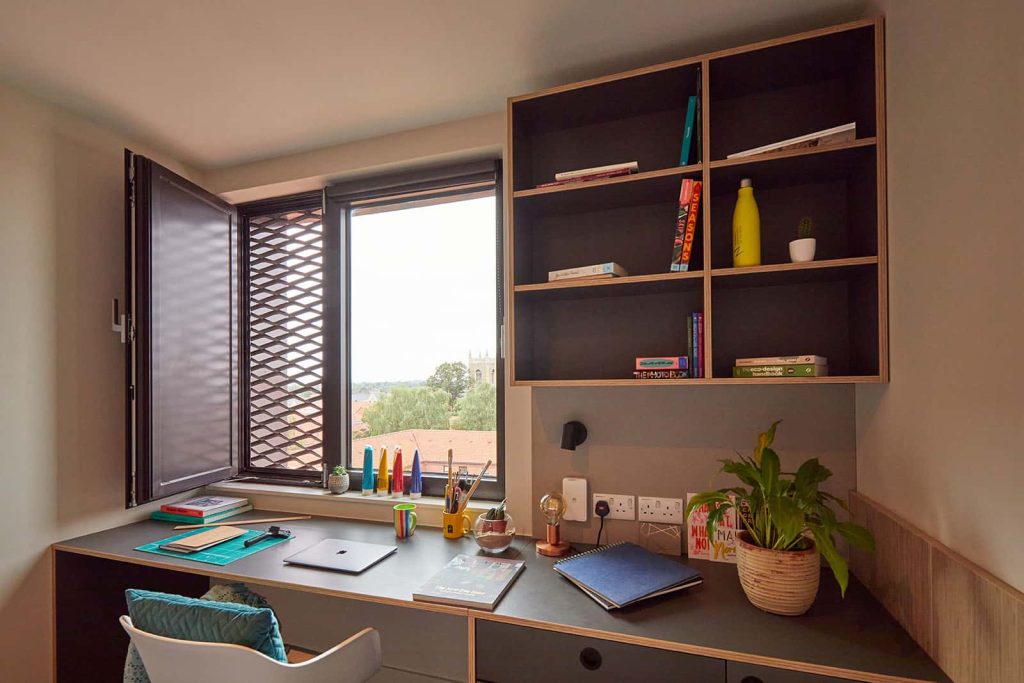 Duke Street Riverside bedroom showing a window open, a laptop on a desk next to a chair with a plant pot, pens, paintings, laptop, shelves and bottle