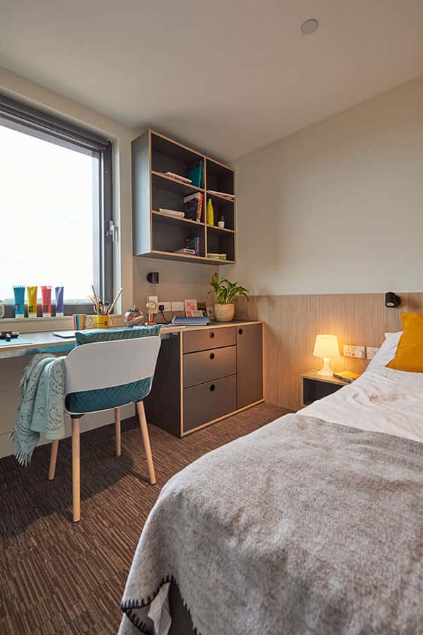 Duke Street Riverside bedroom showing a chair with a throw on, grey throw on bed, colourful paints on a window sill and a shelf full of objects