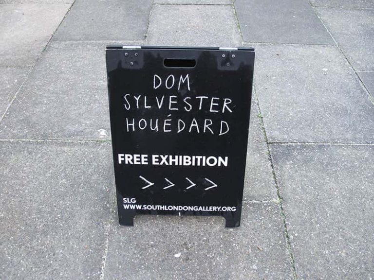 concrete floor with black standing chalkboard in the middle saying Dom Sylvester Houédard, free exhibition. SLG www.southlondongallery.org