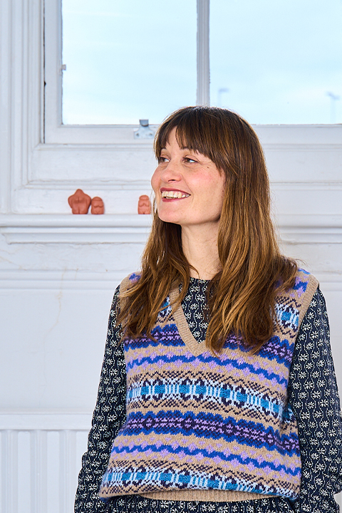 Nicola Simpson stands beneath a window with a white wall behind her. She is wearing a colourful knitted vest and has long brown hair.