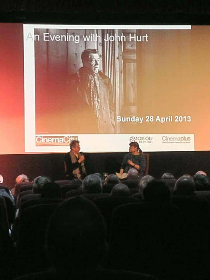 Two people sitting in front of a crowd with a projector screen behind them