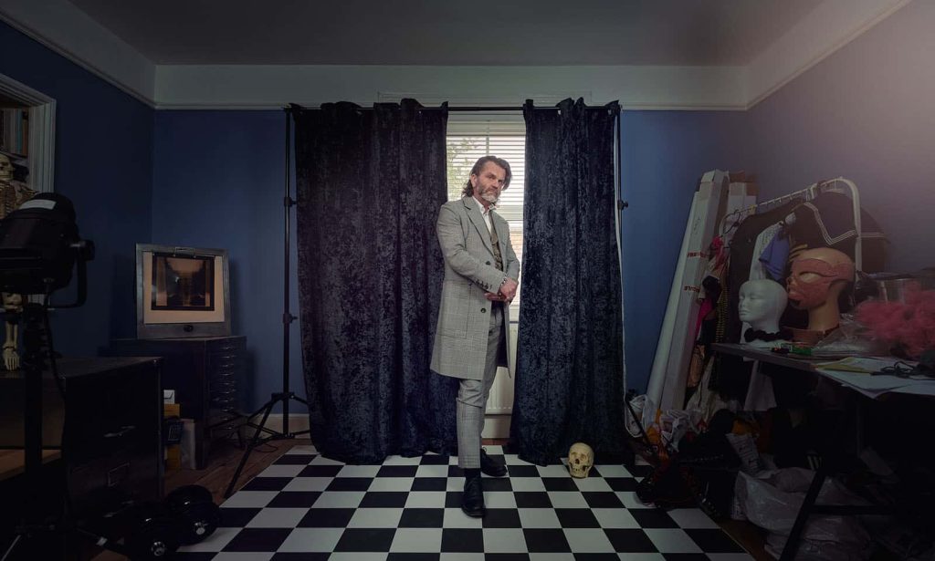 Person standing in dark room with grey suit on standing on checkered flooring. There is a velvet blue curtain behind the person and blue walls in the room. To the left and right of the person there are lots of props such as masks, lights, a clothing rail and a skull.