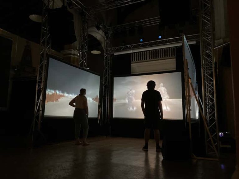 Two people standing in a dark room, in front of three screens with a projection of their bodies on