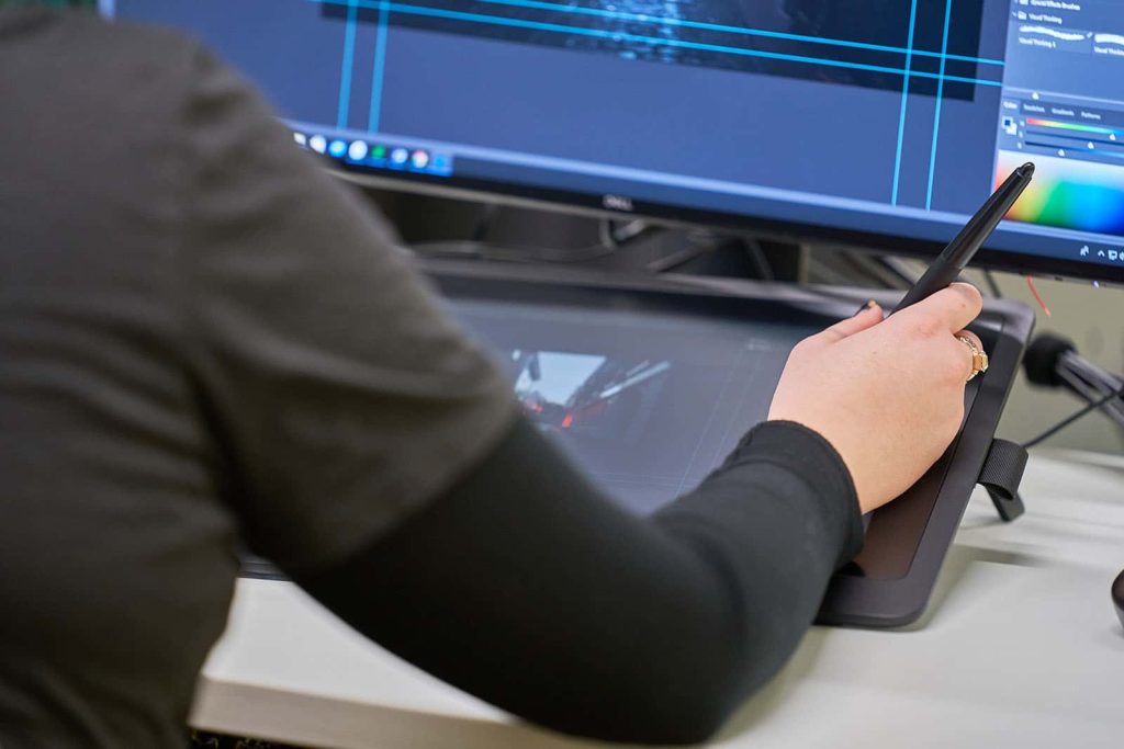 BA Animation student holds a stylus in their hand whilst wearing a black top next to a tablet and computer