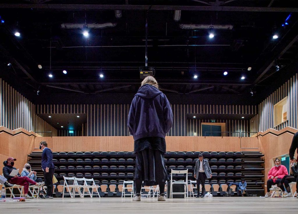 A student in baggy clothes — a baggy hoody, over a long baggy skirt — faces an empty seating area from the centre of a stage, under spotlights on a high ceiling.