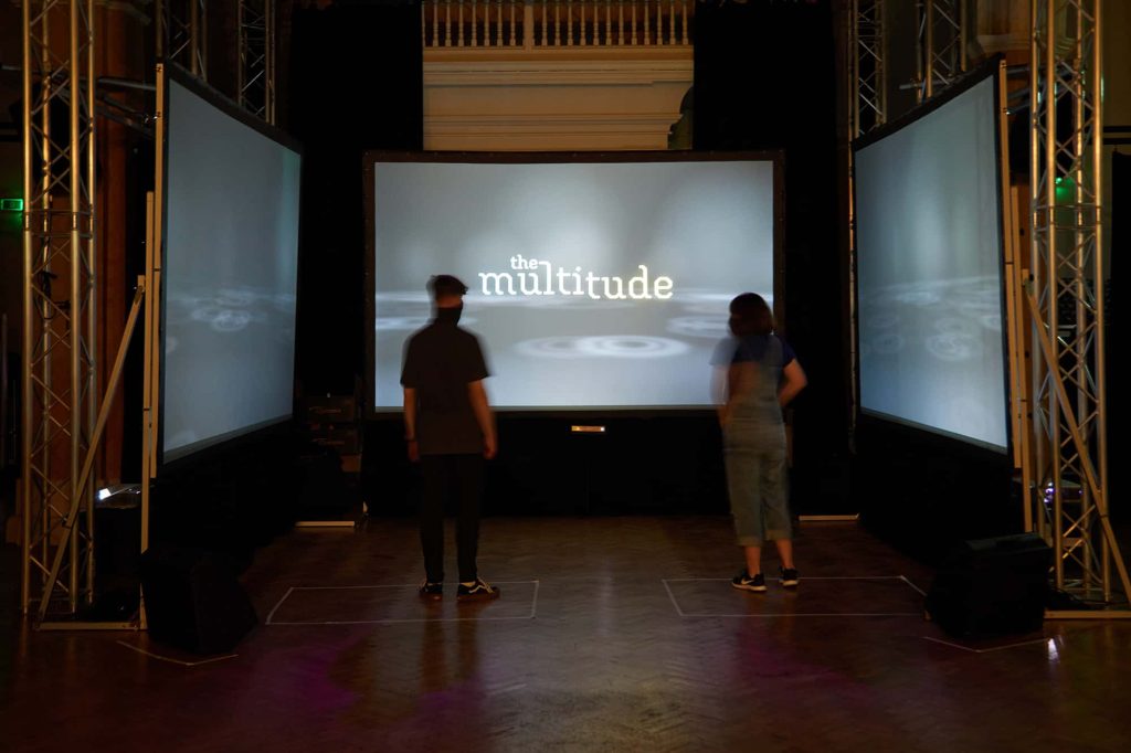 Two people stand in front of 3 screens standing waiting for an instruction from a digital artwork