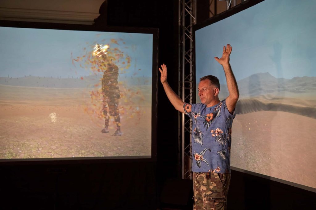 Jamie Gledhill stands in front of two screens in shorts and a t-shirt