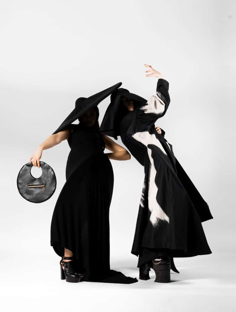 Two womenswear models dressed in avant-garde style black garments, with flashes of white fabric. The models are posed in angular stances. By Emily Rose, BA Fashion