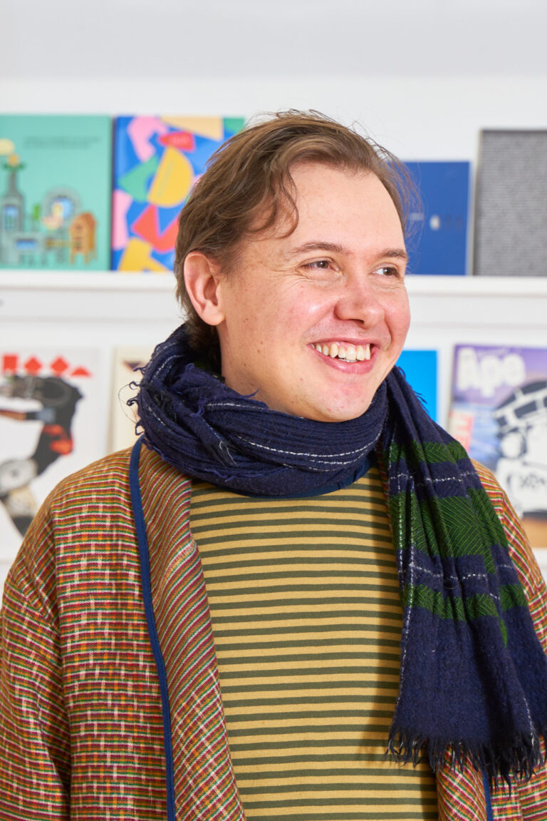 Illustration lecturer Christian Petersen smiles in front of a showcase of student work at Norwich University of the Arts. Christian wears a navy blue scarf with green checks, yellow and grey striped shirt, and a colourful woven cardigan