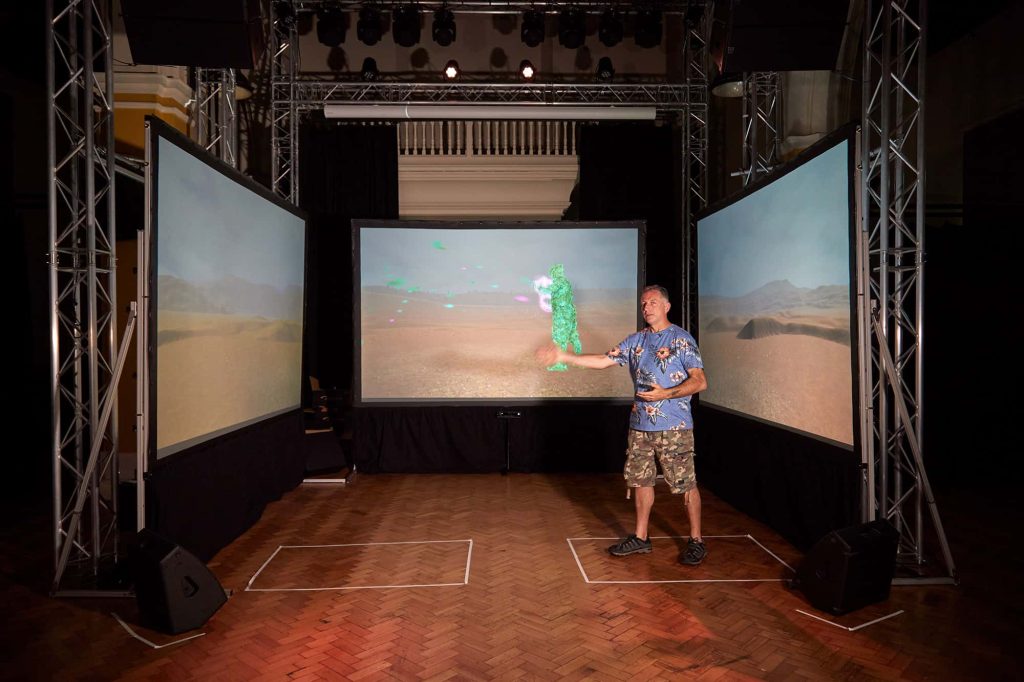 Jamie Gledhill stands in front of three screens wearing shorts and a shirt in front of a game