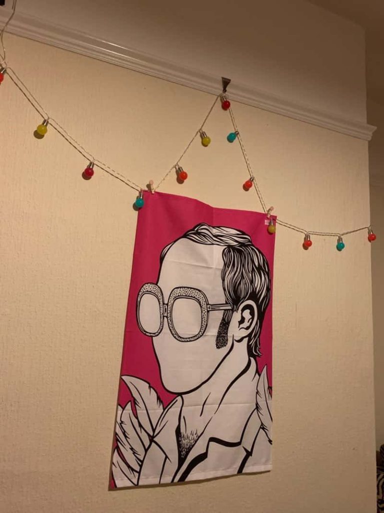 A wall hanging of musician Elton John in the style of artist Julian Opie; a black outline portrait filled in white with minimal detail. Multicoloured lights are draped on the wall above the hanging.