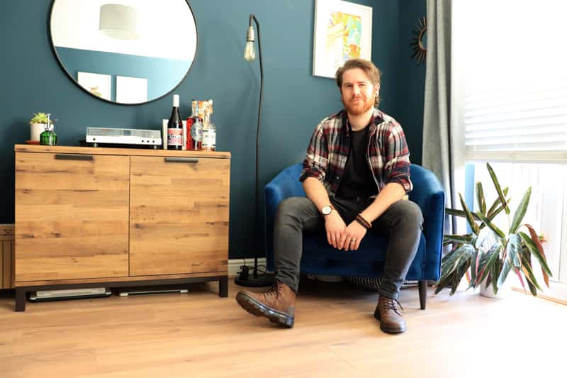 BA Graphic Design graduate Connor Edwards sat on a blue velvet chair in his home, looking at the camera. Next to him on a sideboard is a selection of his alcohol packaging designs