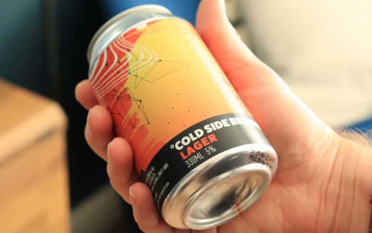 Close up photo of a beer can design for Virgin Wines' own brand 'Cold Side Brewing'. The label shows an abstract orange liquified pattern with thin black linear illustrations. The bottom of the label is solid black, with the branding for Cold Side Brewing, which has a degree symbol before the C in 'Cold'.