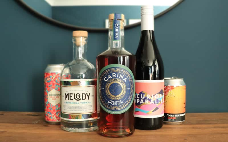 A row of alcoholic drinks placed on a wooden sideboard against a dark blue wall. From left to right, a can of Kaleidoscope beer, with geometric multicoloured pattern on the label. Next, a bottle of clear 'Melody' vodka, with a holographic botanical print pattern and the words 'Melody'. In the centre, a bottle of golden pineapple spiced Carina rum, which has a circular blue label with gold foiling in the pattern of an astrolabe. Next, a bottle of red wine called 'Curious Parallel', with a flat blocky print label of mountains made from pink, purple and turquoise mountains with flashes of gold foil. The words 'Curious Parallel' are juxtaposed amongst the mountains. Finally, a small can of Cold Side Brewing lager, with a vibrant orange label with linear scientific-inspired pattern.