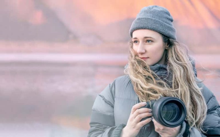 A photograph of BA Photography graduate Cat Gundry-Beck holding a DSLR camera, wearing a grey puffer jacket and grey beanie hat, looking beyond the camera