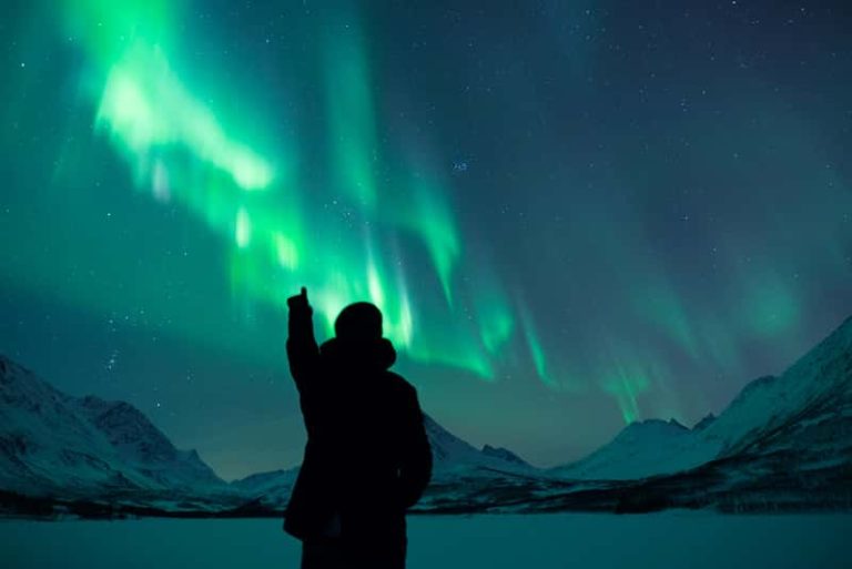 A landscape photograph of a silhouette pointing up at the aurora borealis (northern lights) in Norway