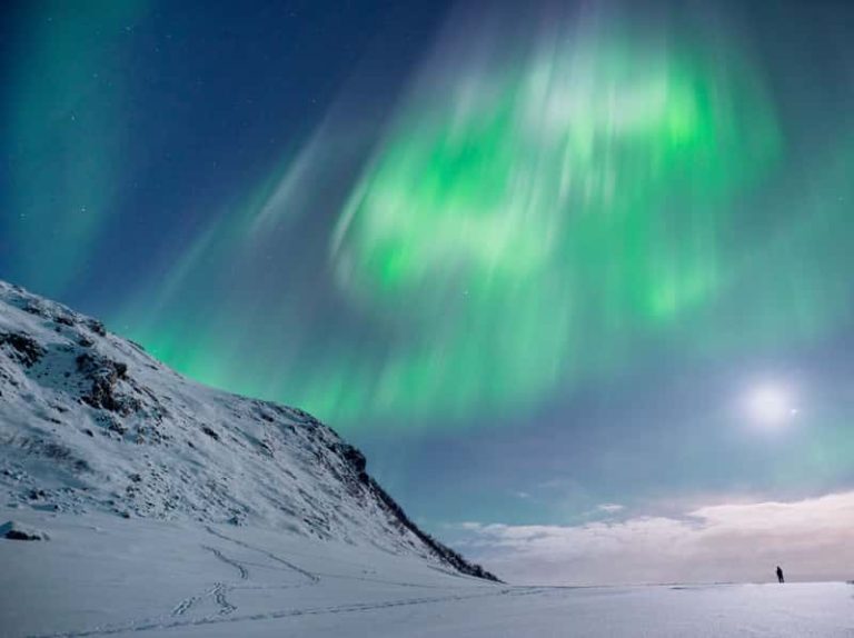 A wide angle landscape photo of a snowy mountain, with a distant silhouette. Above the mountain is the aurora borealis in vibrant green and white colours, against an inky blue sky filled with stars