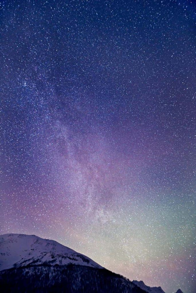 A portrait photograph of the night sky with the top of a mountain in the foreground. The sky fades from inky blue, to purple, to green, where the aurora borealis filters in amongst the stars