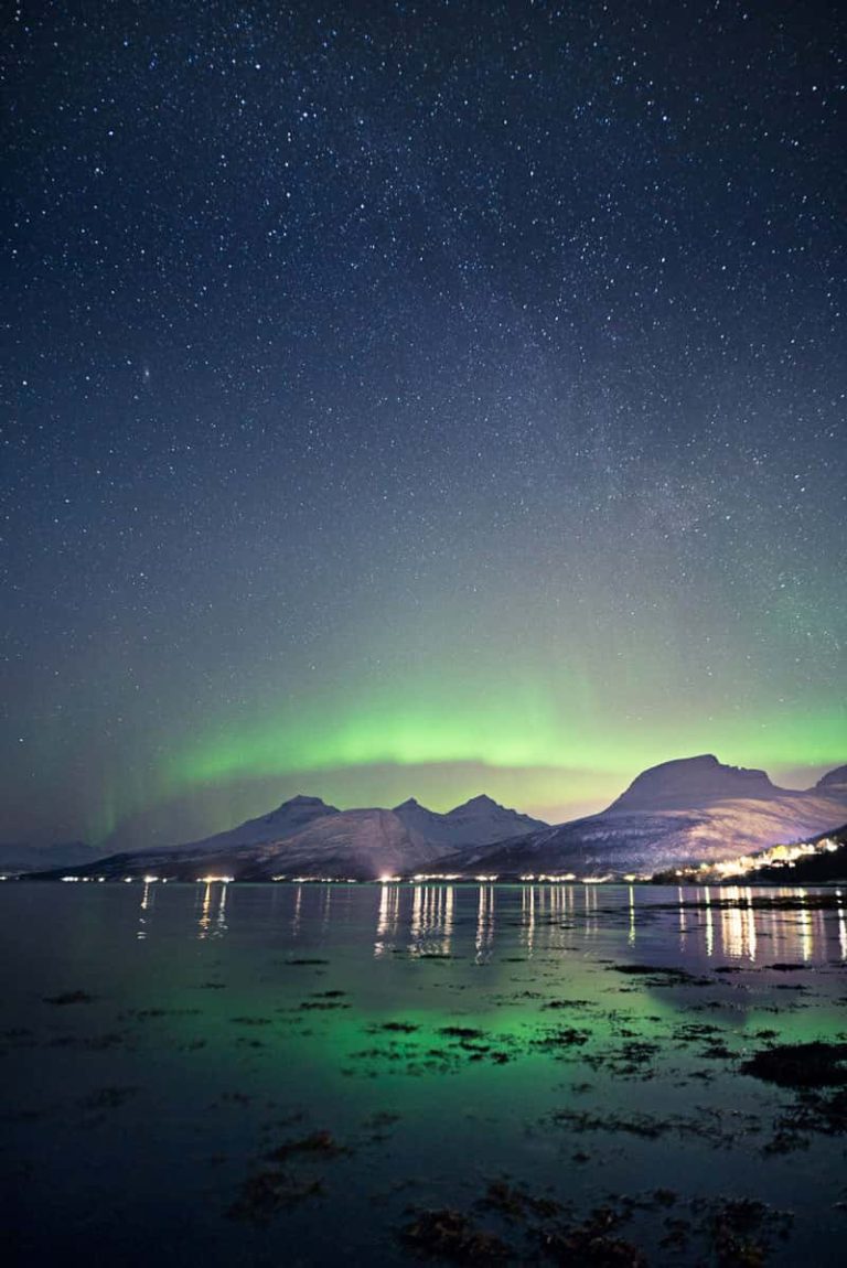 A wide angle portrait photo of distant mountains, lit up in shades of purple, with the green hues of the aurora borealis hovering above the peaks. The sky is littered with stars, fading from the aurora green into deeper shades of inky blue. In the foreground, a mass of water reflecting the sky.