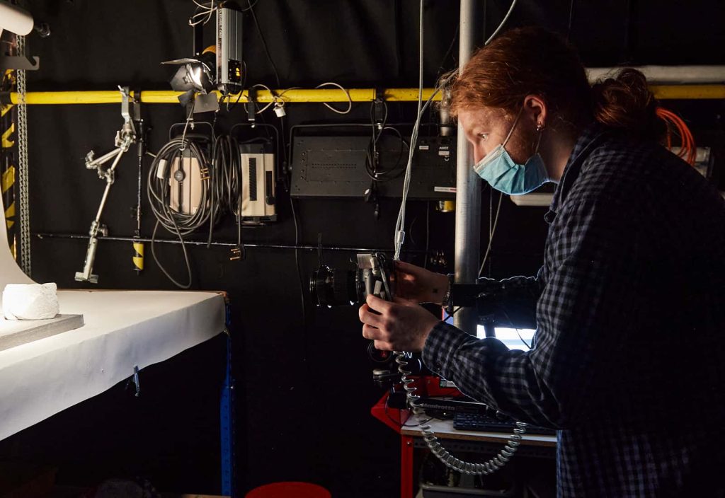 A student leans in to shoot their project in the animation studio. They have long ginger hair, and wear a dark blue chequered shirt. In the background, there is lots of equipment and hardware hung on the wall.