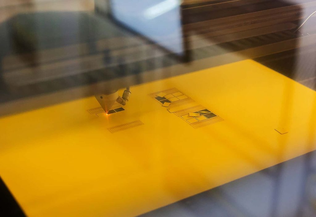 One of the laser cutters at Norwich University of the Arts cuts the small intricate shapes of wooden window shutters out of a piece of yellow card