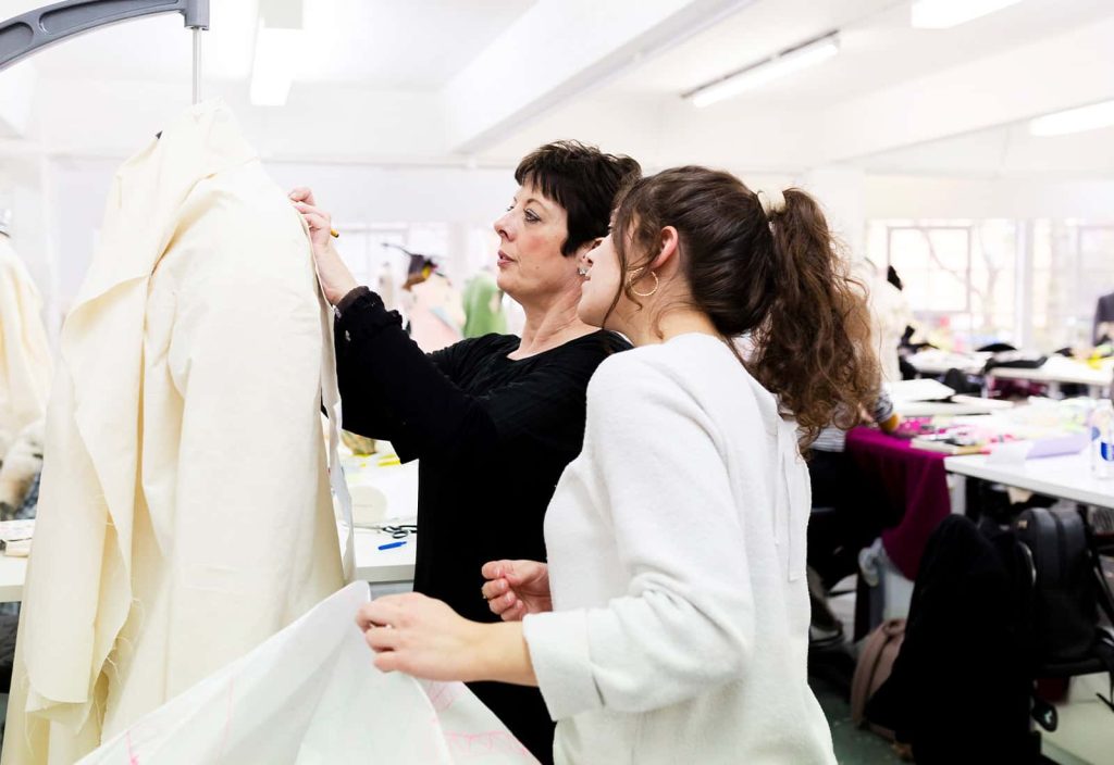 Sue Chowles teaches a student in the fashion workshop. The student is holding a pattern and watching Sue mark fabric which she's holding up to a mannequins shoulder