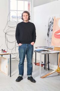 Alex Crocker standing in the Fine Art studio, wearing a black sweatshirt, blue jeans, and black doc martens